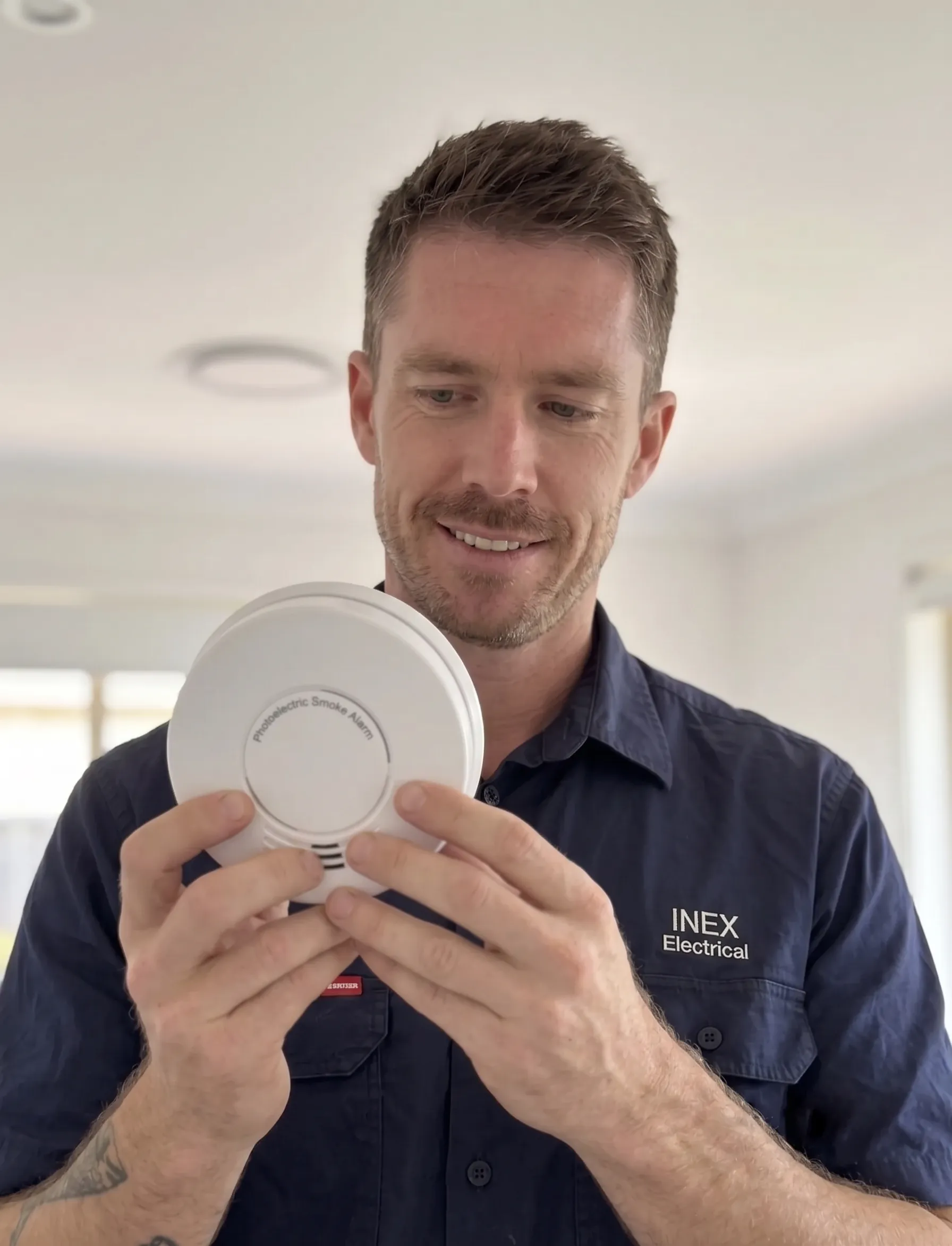 James, an electrician from INEX Electrical servicing the Gold Coast, conducting a residential compliance check. James is holding a modern, clean white photoelectric smoke alarm.